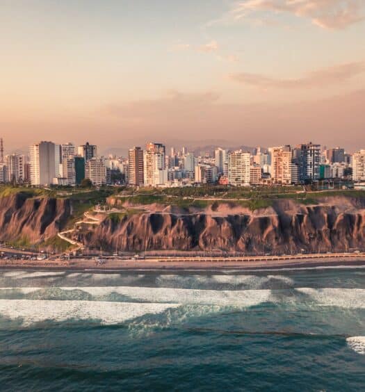 Imagem da Playa de Costa Verde em Lima, é possível ver o mar, uma faixa de areia, logo em seguida, uma formação rochosa e após isso, uma avenida e a cidade ao fundo