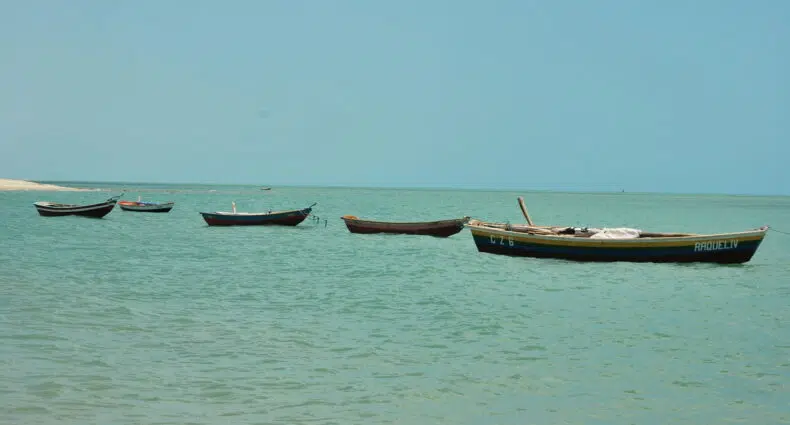 Quatro pequenos barcos estão flutuando em águas turquesas calmas sob um céu azul claro perto de uma costa arenosa, em Barra Grande no Piauí, para representar onde ficar na Barra Grande no Piauí.