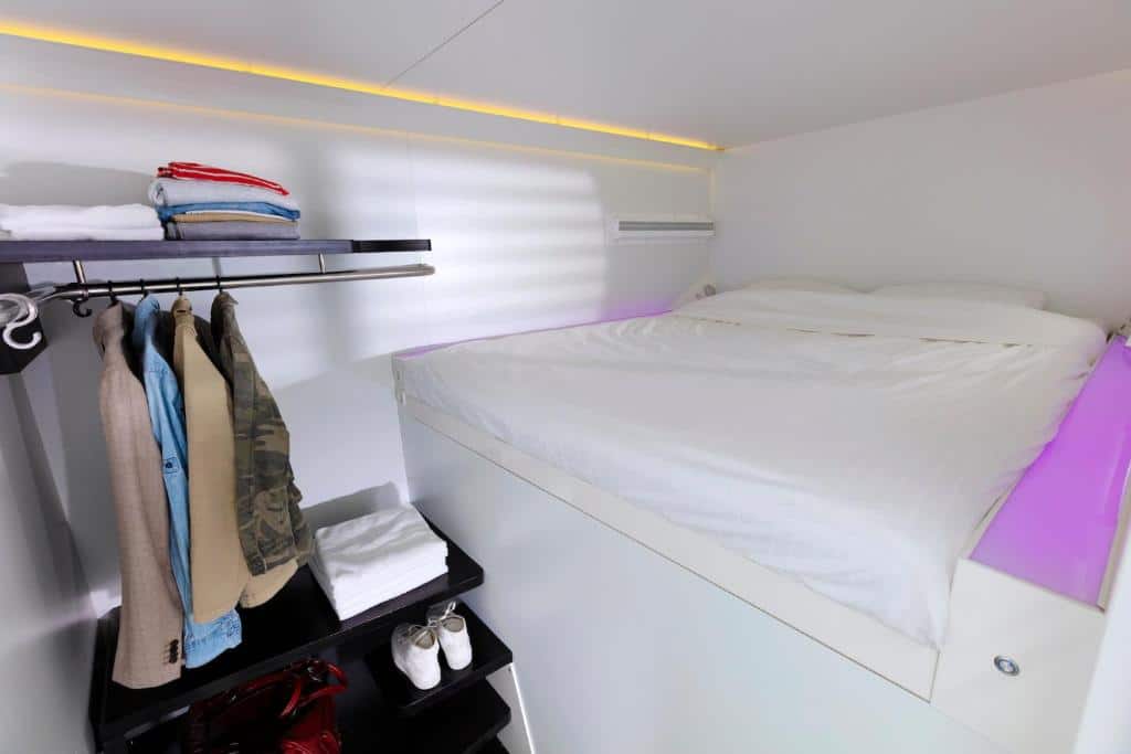 Interior de uma das capsulas do CityHub Amsterdam, de 6 m², com um pequeno cabideiro e prateleiras ao lado com roupas dobradas e sapatos, e colchão com travesseiros e ar-condicionado