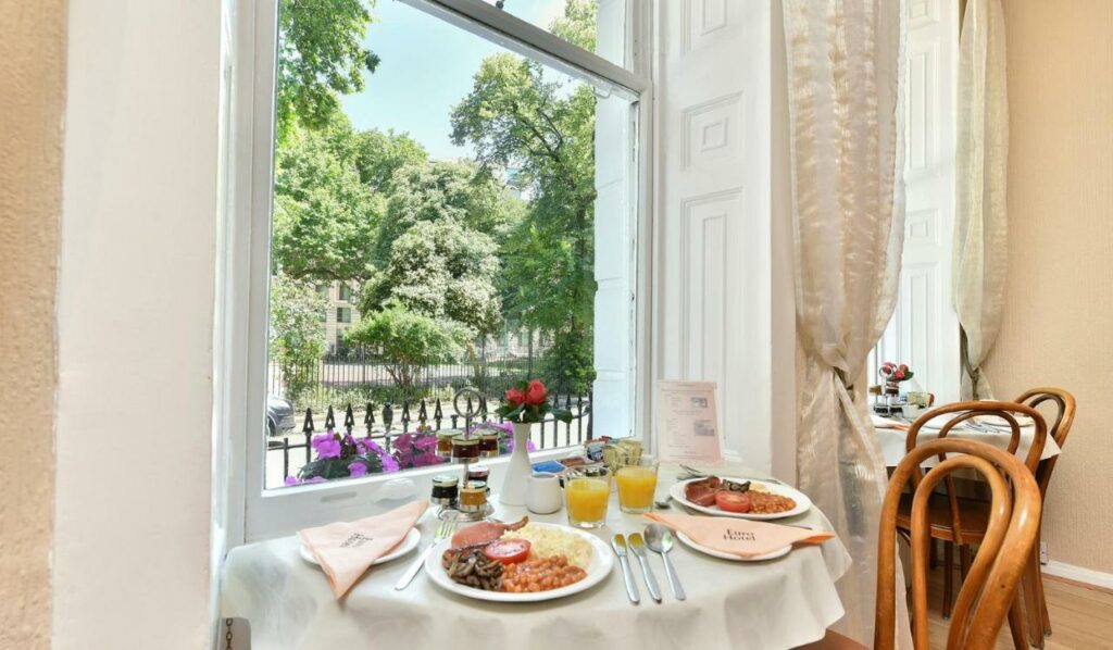 Mesa do restaurante do Euro Hotel virada para uma ampla janela com vista para a rua, árvores e flores, sob a mesa uma refeição completa e tudo decorado de forma delicada