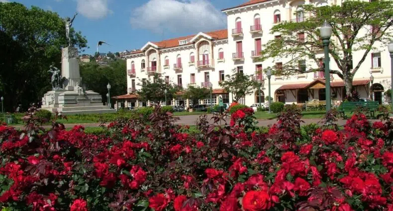 Rosas vermelhas em primeiro plano, com um parque, uma estátua e um grande edifício branco do Palace Hotel - Poços de Caldas, uma das opções entre as pousadas em Poços de Caldas, ao fundo, sob um céu azul.