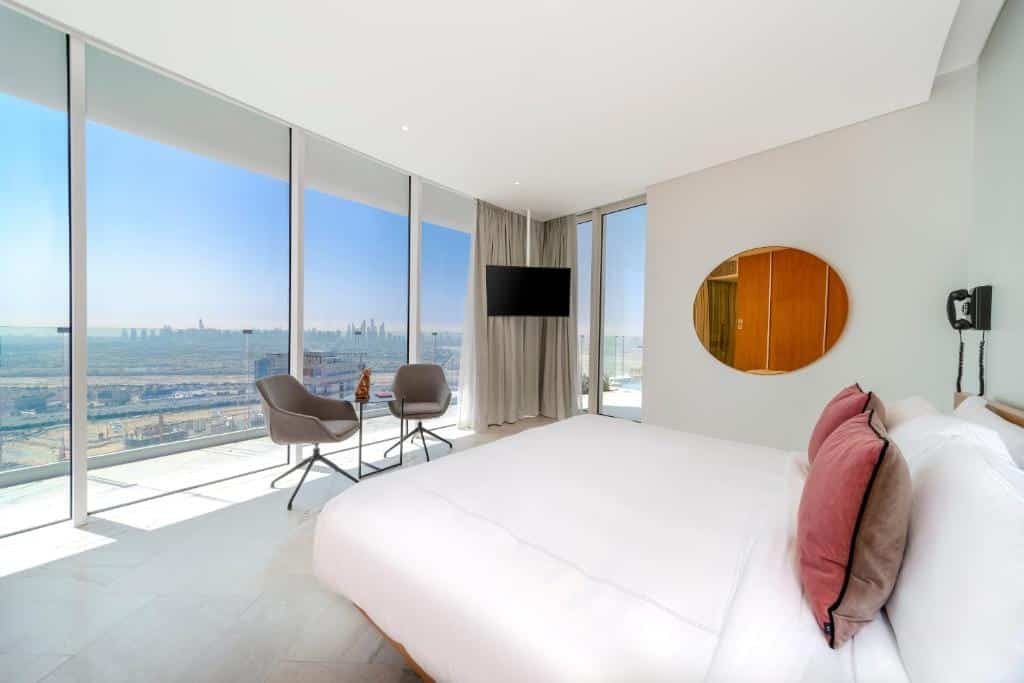 Quarto do Five Jumeirah Village, um dos hotéis em Dubai, com cama, duas cadeiras, espelho, TV e janelas de vidro com vista da cidade