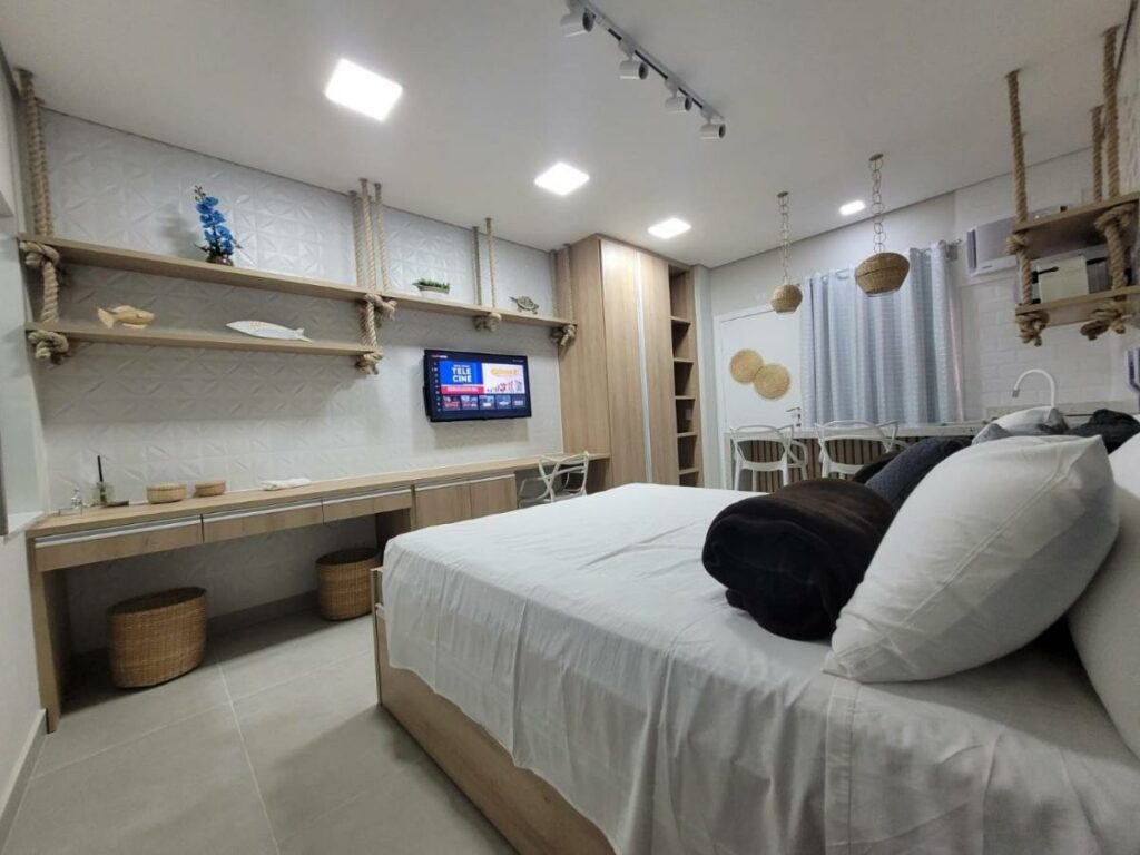 Quarto do Flat Amarilis Apartamento 103, com decoração praiana, uma cama de casal, uma bancada com duas cadeiras e uma pequena cozinha, iluminação indireta próxima da cama e uma pequeno armário