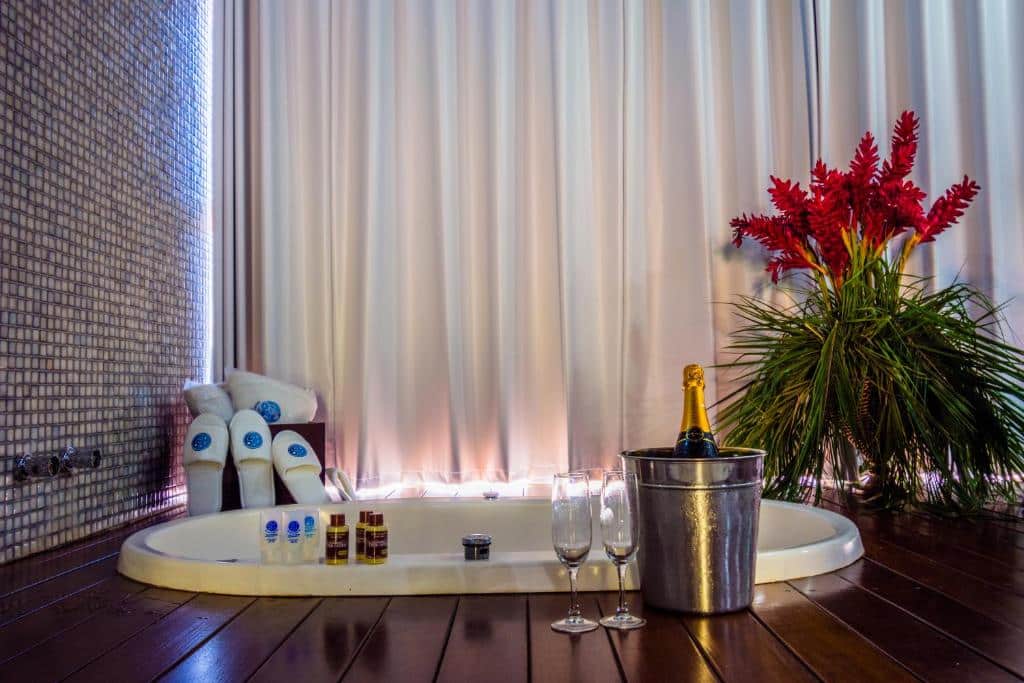 Hidromassagem no Balneário do Lago Hotel, com um deck ao redor, taças, um vinho, alguns acessórios de banho, um vaso de flor e uma janela com cortinas