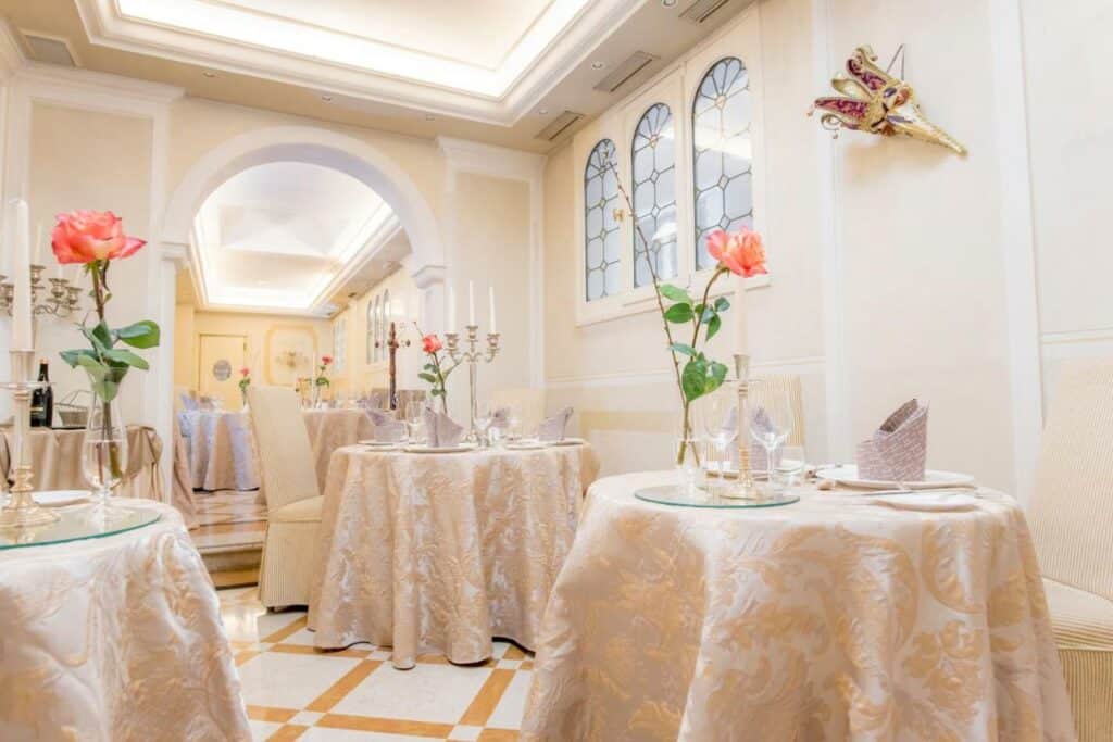 Restaurante do Hotel Carlton On The Grand Canal  com mesas redondas e toalhas bordadas, com flores e taças sob as mesas, ambiente em tons de bege claro e branco, para representar hotéis em Veneza
