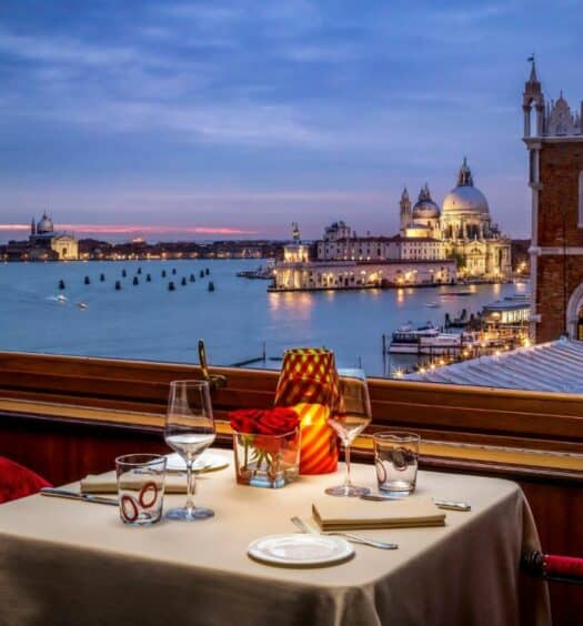 Varanda do Hotel Danieli com uma vista noturna do canal de Veneza com tudo iluminado e uma mesa com dois lugares com taças e velas