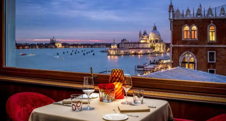 Varanda do Hotel Danieli com uma vista noturna do canal de Veneza com tudo iluminado e uma mesa com dois lugares com taças e velas
