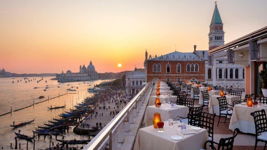 Varanda do Hotel Danieli, em Veneza, com pôr do sol alaranjado, várias mesas espalhadas e vista do mar com caiaques enfileirados