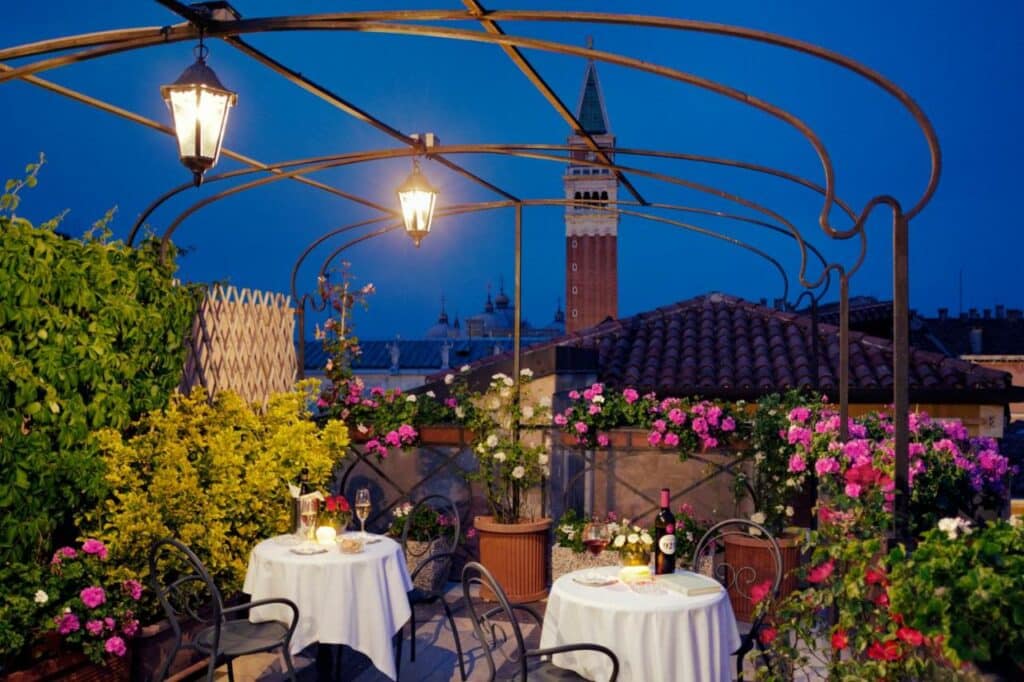 Terraço do Hotel Firenze cercado por flores rosas e vermelhas, duas mesas redondas com dois lugares, com taças e velas sob as mesas, para representar hotéis em Veneza