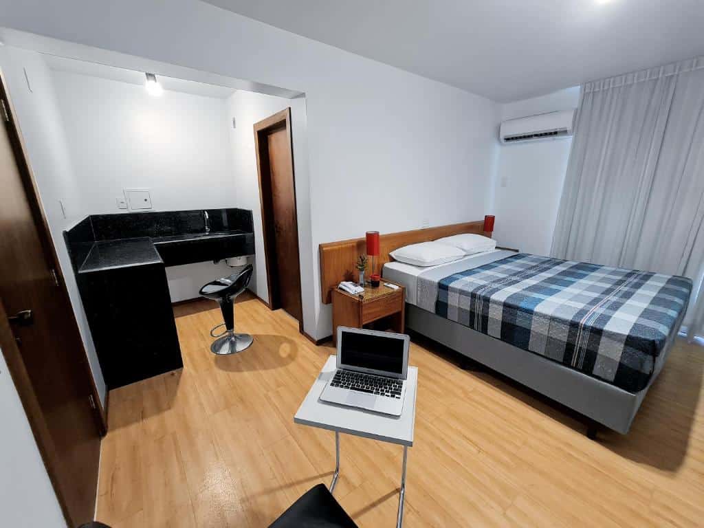 Quarto do Germânia, com uma cama de casal, mesa pequena com computador e balcão ao lado da porta do banheiro
