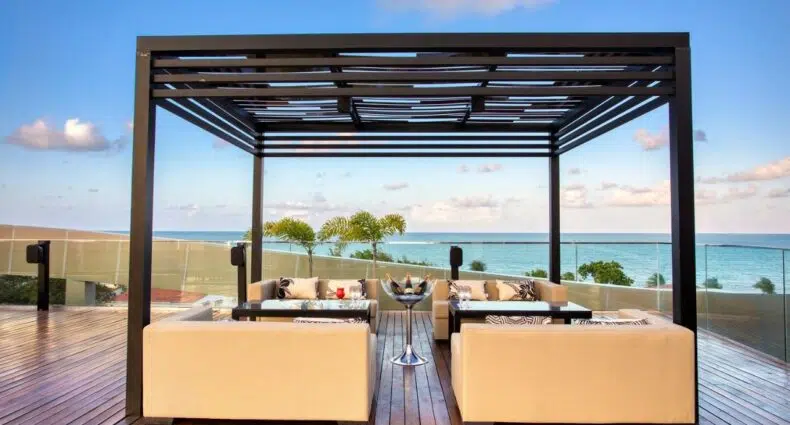 Área de lounge externa com sofás bege sob uma pérgola em um deck de madeira, com vista para o oceano e o céu azul.