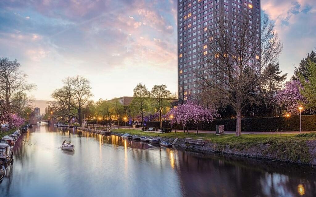 Prédio do Hotel Okura Amsterdam, um dos hotéis de luxo, com um canal grande com as águas refletindo as luzes do prédio e casas ao lado. Há também árvores com flores roxas a frente