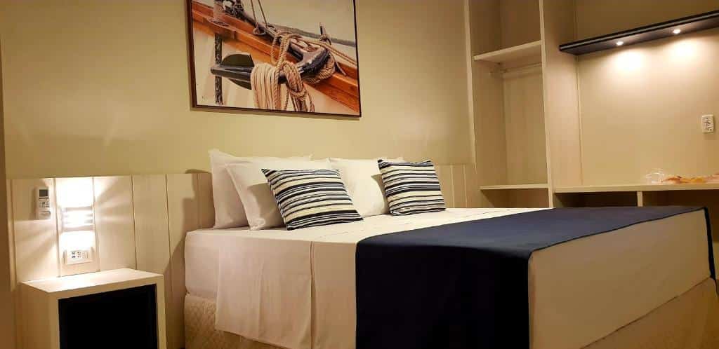 Quarto do Hotel Pousada Paradise, em Caraguá, com cama de casal, mesa de cabeceira, quadro decorativo com imagem de 
âncora e cordas de marinheiro, e roupa de cama branca e azul marinho