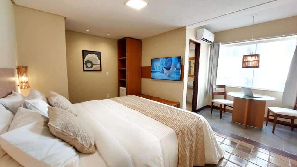 Quarto no Kastel Jampa com uma cama de casal, televisão, mesa com dois lugares, decoração com detalhes em bambu, um armário de conceito aberto, ar-condicionado e uma janela, para representar resorts em João Pessoa