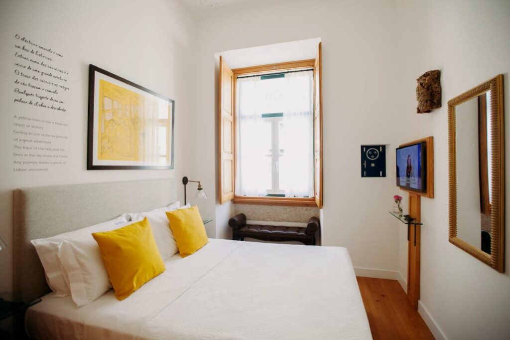 Quarto de casal no Otília Apartamentos, com almofada amarela e quadro amarelo na parede, espelho em frente à cama e ampla janela na parede ao fundo