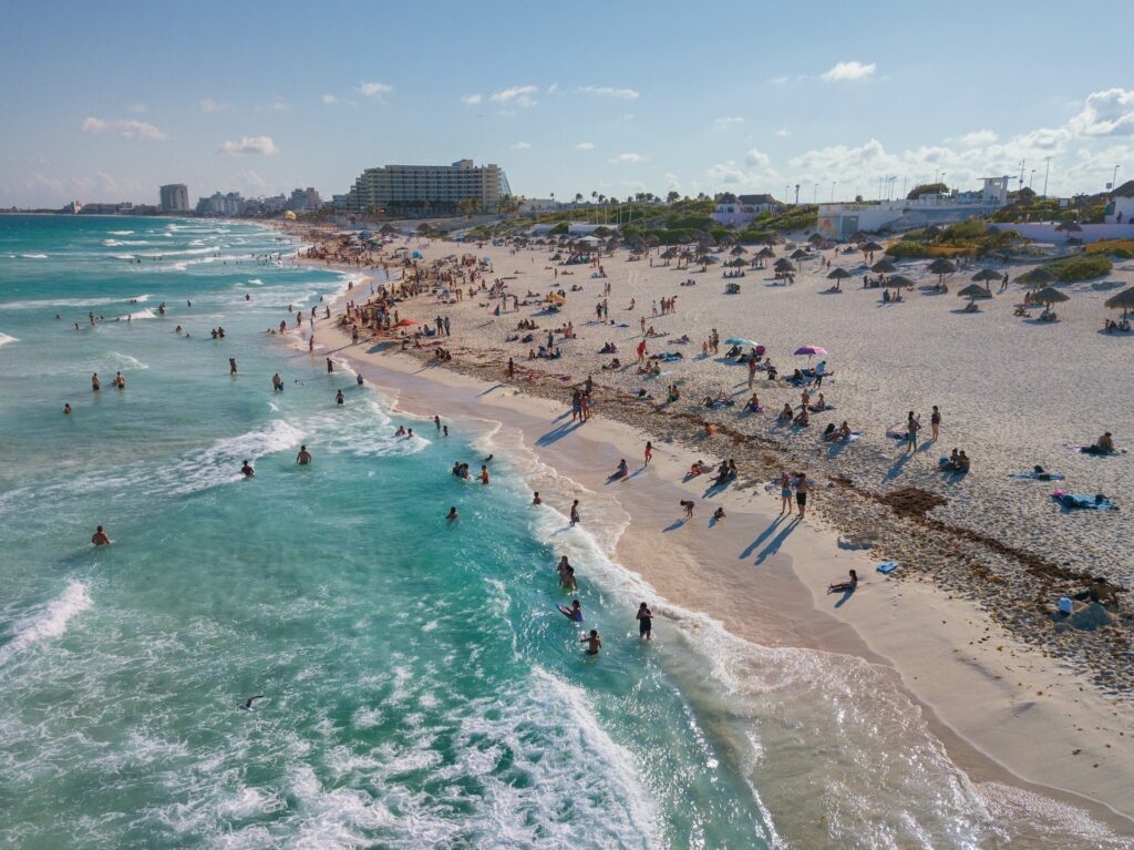 Playa Nizuc na Zona Hoteleira em Cancun, com uma extensa orla, faixa de areia longa e o mar de água verde clara, com muitas pessoas, e alguns hotéis logo depois da praia
