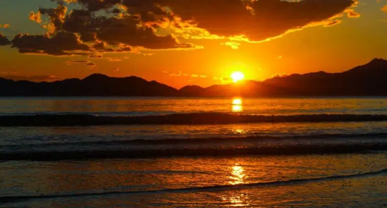 Pôr do sol sobre montanhas distantes com céu laranja, nuvens e luz do sol refletindo nas ondas calmas do oceano em primeiro plano para ilustrar o post de pousadas em Bertioga