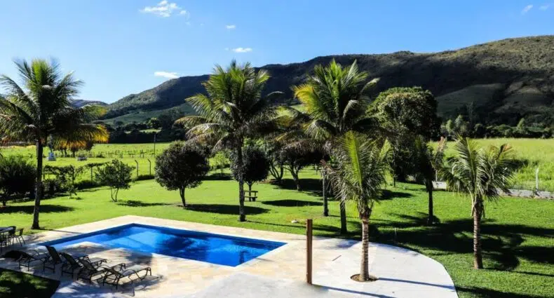 Piscina retangular cercada por espreguiçadeiras, palmeiras e campos gramados com colinas ao fundo sob um céu azul. Essa é a Pousada Cateto - Zona Rural, uma das pousadas em Capitólio.