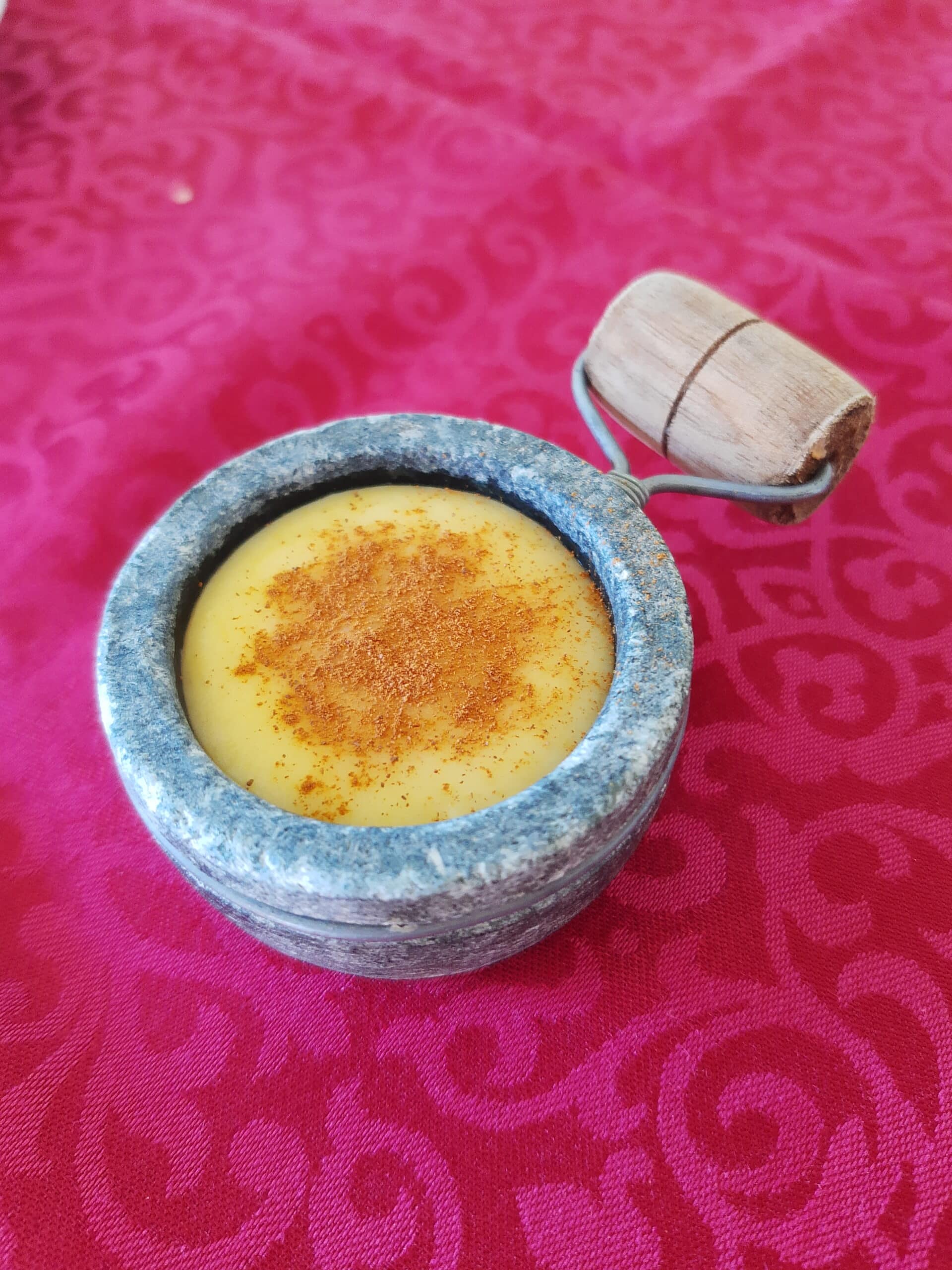 potinho de pedra sabão com curau de milho com canela em pó em cima de uma toalha de mesa vermelha
