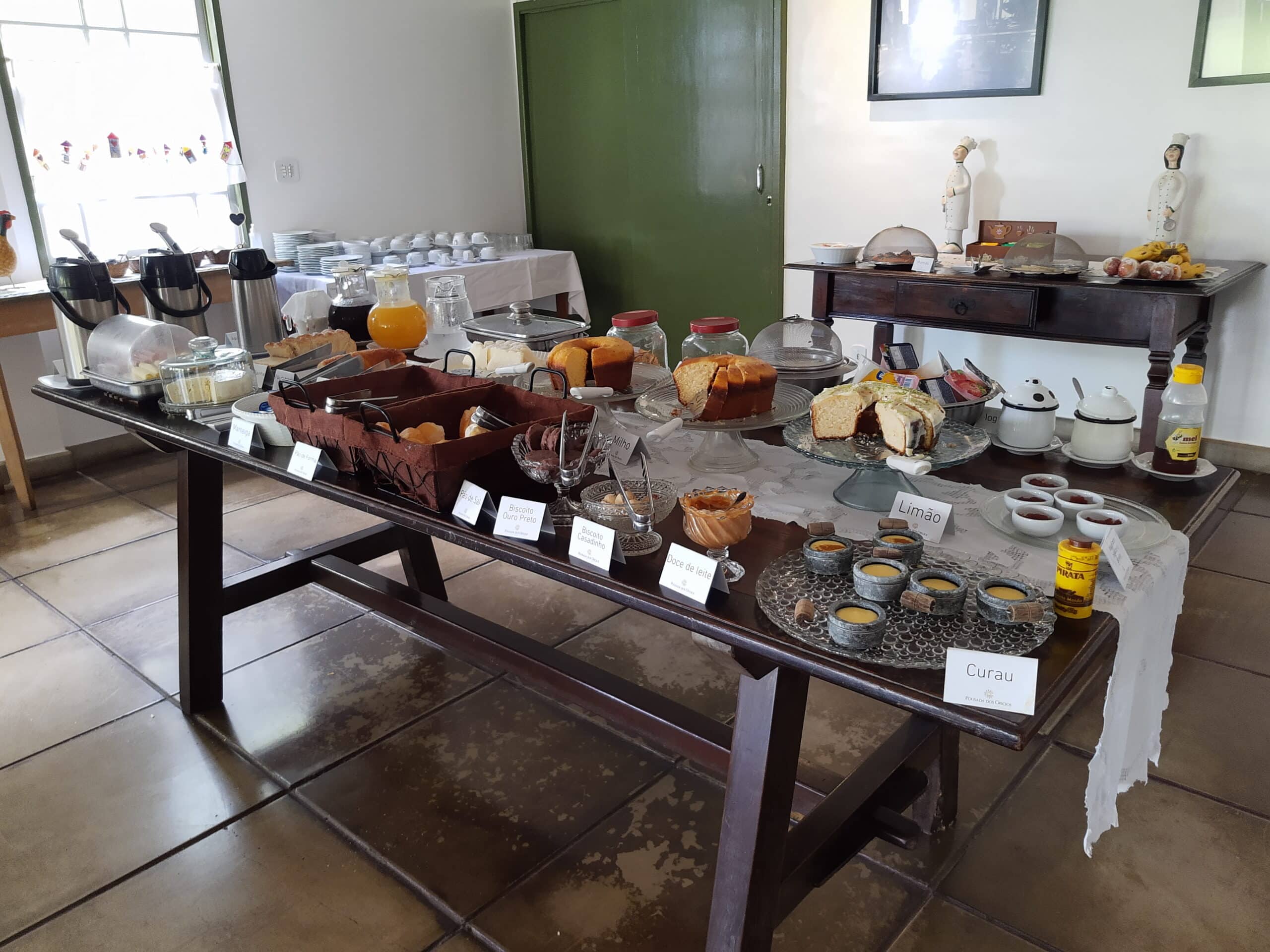 mesa farta de alimentos, como pães, bolos, cereais, bandejas de frios, sucos e ovos mexidos na área de café da manhã da pousada dos ofícios
