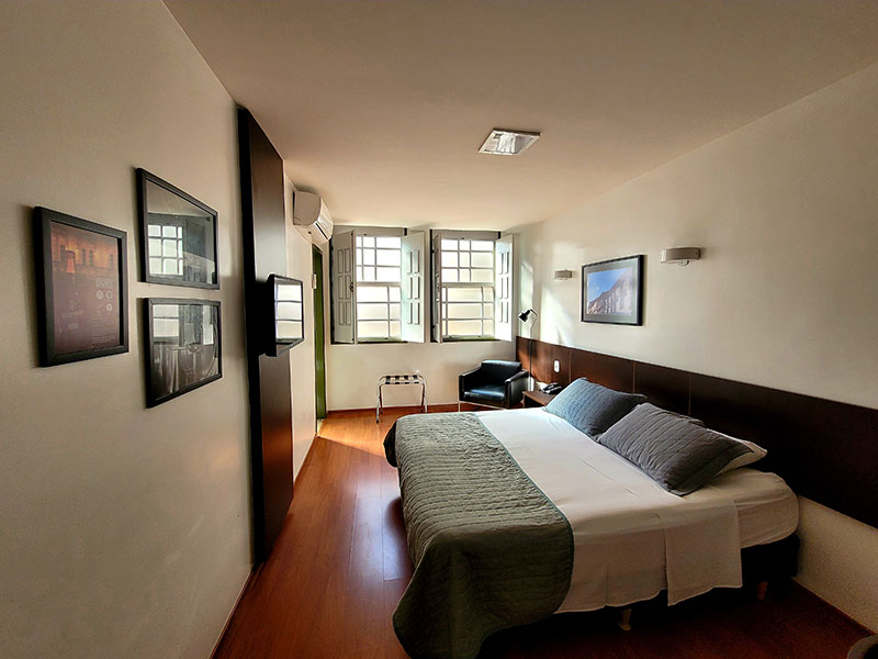 quarto superior com uma cama de casal, duas janelas abertas, um ar-condicionado, uma TV de tela plana e alguns quadros decorativos