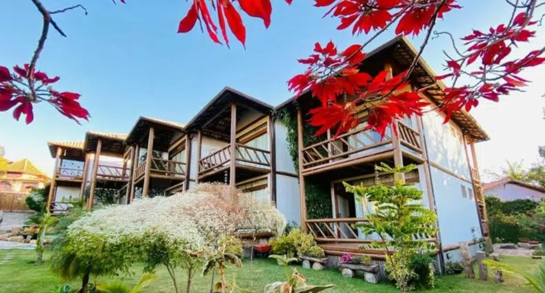 Edifício de dois andares com varandas de madeira, cercado por plantas verdes e folhas vermelhas em primeiro plano.