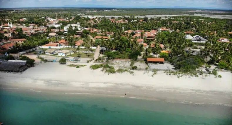 Vista aérea da Na Praia Pousada, uma das pousadas em Barra Grande no Piauí com praia de areia, árvores verdes e casas próximas à costa sob um céu parcialmente nublado.