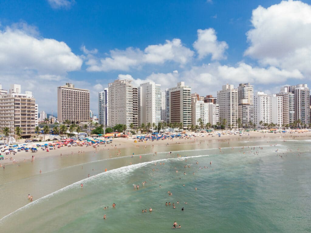 Praia das Pitangueiras no Guarujá, com uma orla extensa, muitas pessoas na água e na areia e os prédios logo atrás