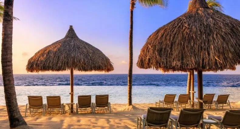 Várias espreguiçadeiras ficam de frente para o oceano sob dois grandes guarda-sóis de palha e palmeiras em uma praia de areia ao pôr do sol no Renaissance Wind Creek Curaçao para ilustrar post sobre resorts em Curaçao.