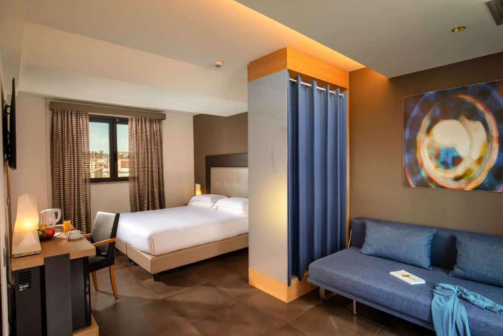 quarto do Best Western Plus Hotel Spring House, um dos hotéis para brasileiros em Roma, com cama de casal e área de estar com sofá baixo azul, há uma mesa e cadeira à frente, com uma janela com cortina