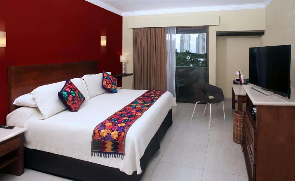 Quarto no Adhara Hacienda Cancun com uma cama de casal, uma mesa com uma cadeira, uma varanda, uma cômoda, uma televisão e duas mesinhas de cabeceira com abajures, para representar hotéis em Cancun