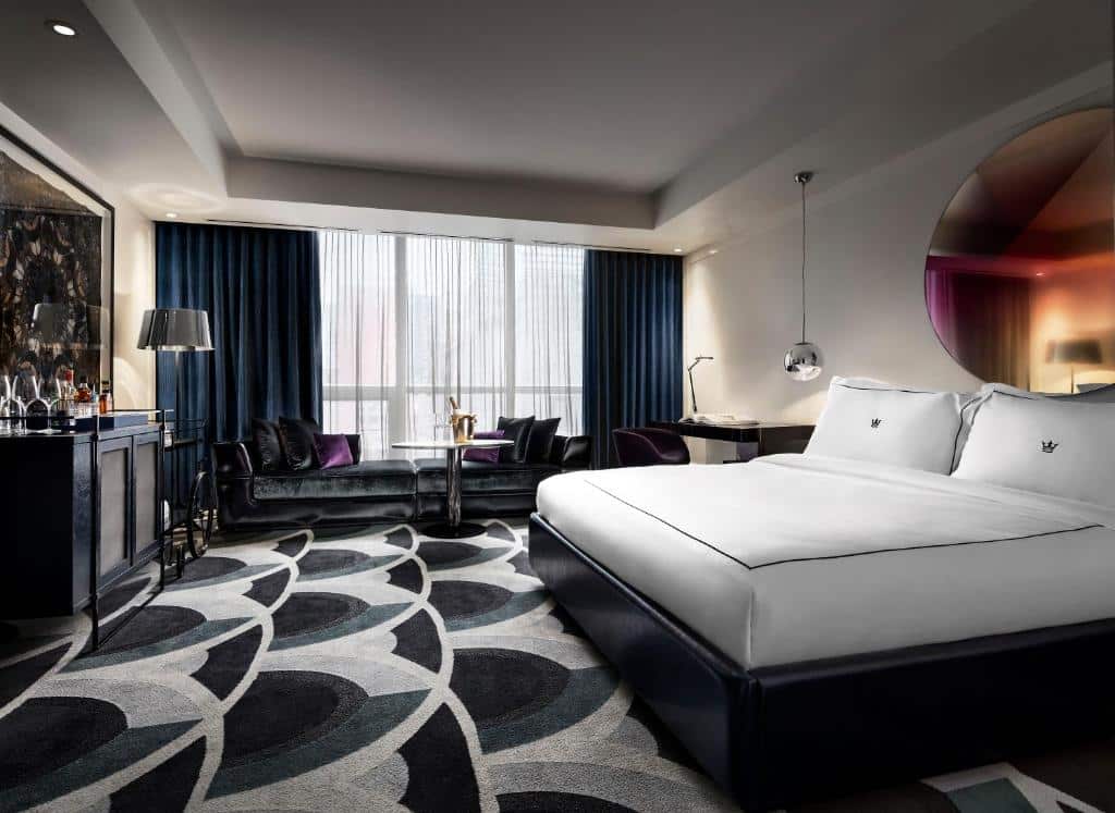 Quarto do Bisha Hotel Toronto, ambiente amplo, decorado em tons de preto, branco e cinza, com uma janela ampla com cortinas, uma mesa e um pequeno sofá com almofadas de veludo, uma mesa de escritório com uma cadeira, uma cama de casal e uma cômoda, para representar hotéis em Toronto