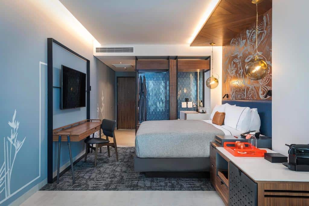 Quarto elegante no Canopy By Hilton Cancun La Isla decorado em tons de cinza e azul, om uma cama de casal, uma mesa com uma cadeira, uma televisão, uma sacada ampla, luminárias suspensas e uma cafeteira