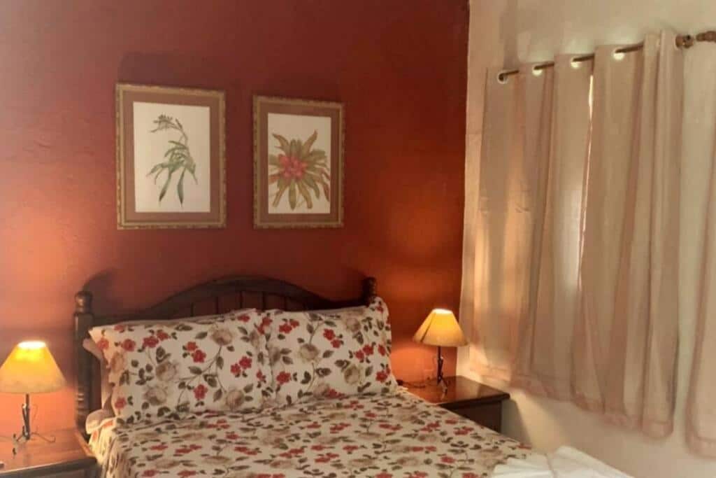 Quarto no Chalé Gaia - Itatiaia com uma cama de casal, uma janela com cortinas, dois abajures, dois quadros e uma parede em tom de vermelho queimado