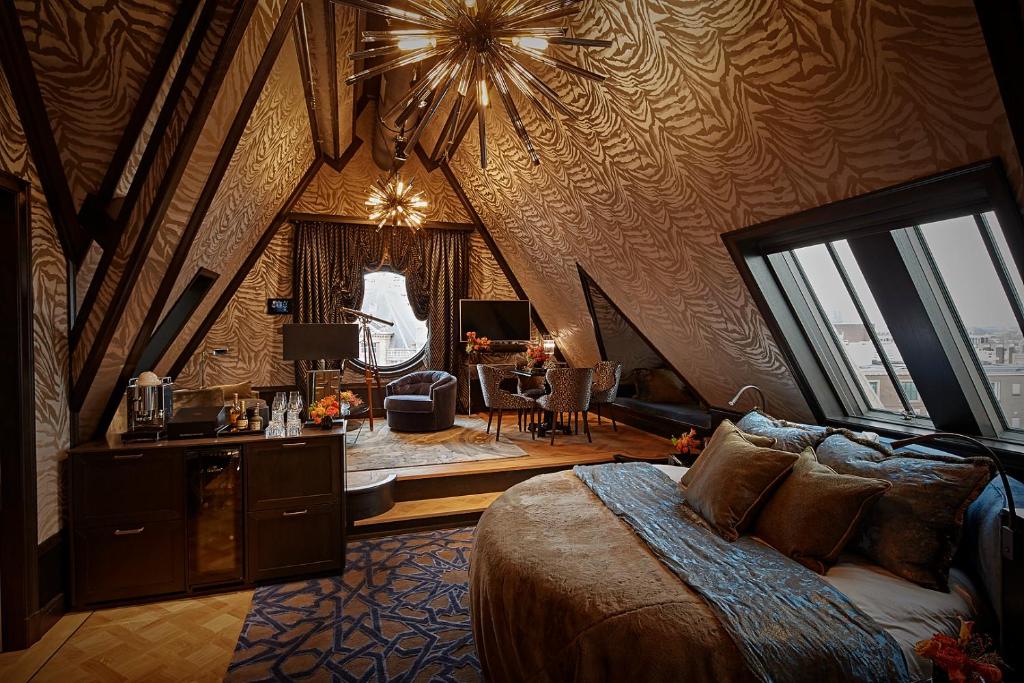 Quarto luxuoso do Hotel TwentySeven, com teto no formato de triangulo com desenhos em curvas num tom de marrom e bege, cama redonda e, ao fundo, uma sala de estar luxuosa com janela redonda e cortina marrom