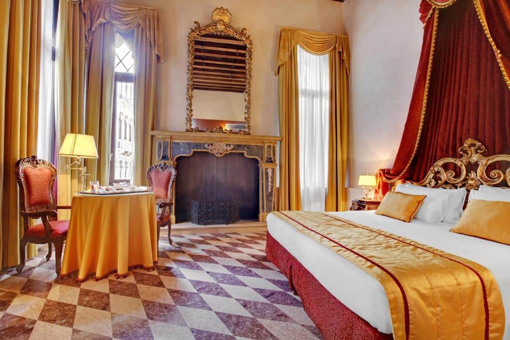Quarto no Hotel Donà Palace, um local amplo com duas janelas, uma lareira, uma cama de casal, uma mesa com duas cadeiras vermelhas, tudo decorado em tons de dourado, vermelho e branco