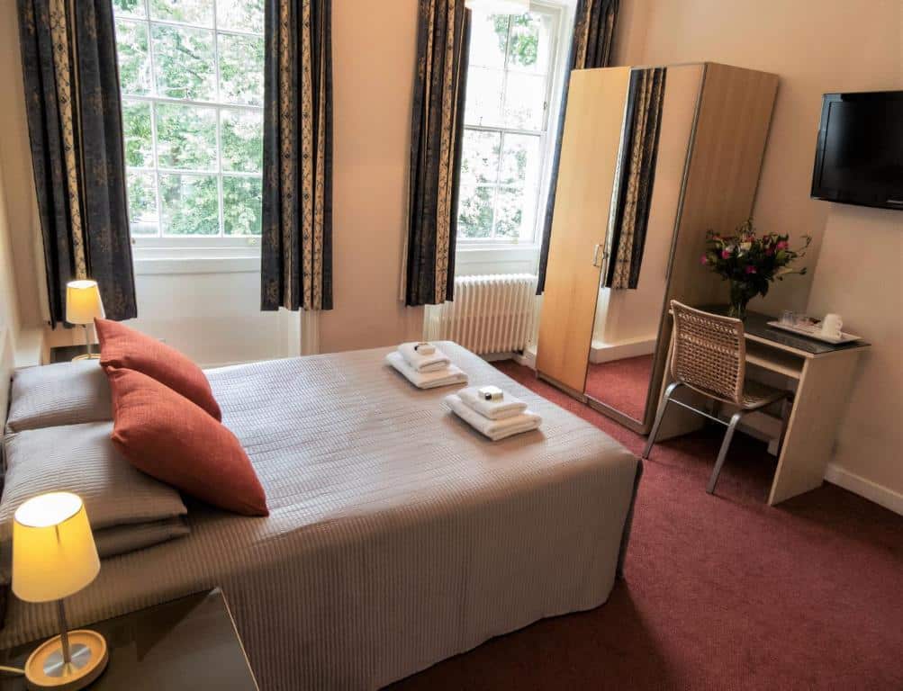 Quarto do Euro Hotel com duas janelas, um pequeno armário com espelho, uma televisão, uma mesa e uma cadeira, e a cama de casal com toalhas e almofadas, para representar hotéis em Londres