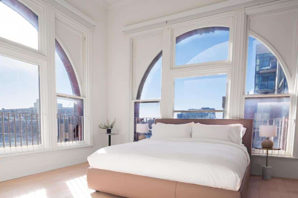 Quarto no Gladstone House com amplas janelas que iluminam o ambiente todo que conta com uma cama, duas mesinhas de cabeceira com abajures, para representar hotéis em Toronto