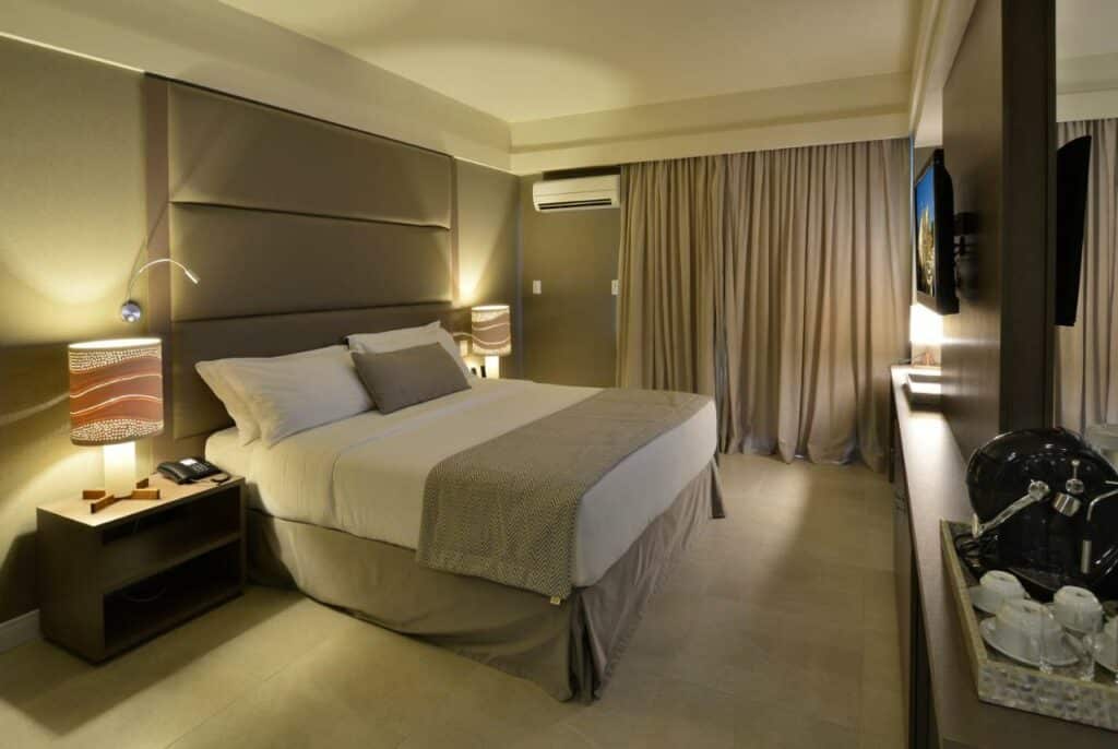 Quarto no Hardman Praia Hotel com uma cama de casal, um ar-condicionado, uma televisão, uma varanda com cortinas e um abajur, para representar resorts em João Pessoa