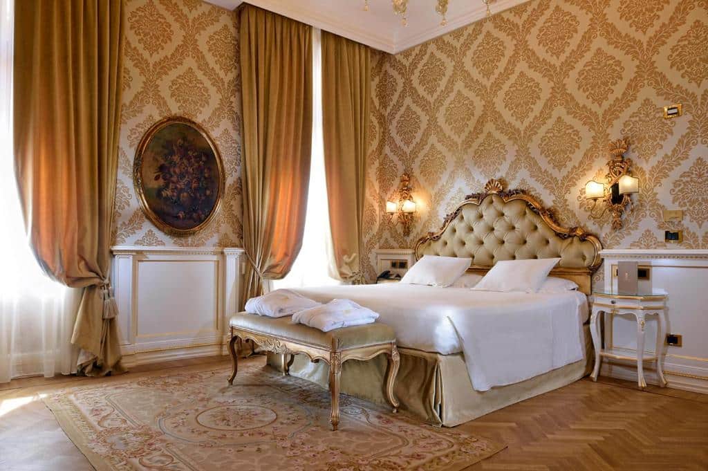 Quarto amplo no Hotel Ai Reali em tons de bege e dourado, decorado em estilo renascentista, com uma cama de casal, uma quadro, duas janelas amplas, uma mesinha de cabeceira e dois abajures presos na parede, chão de madeira e um tapete bege, para representar hotéis em Veneza
