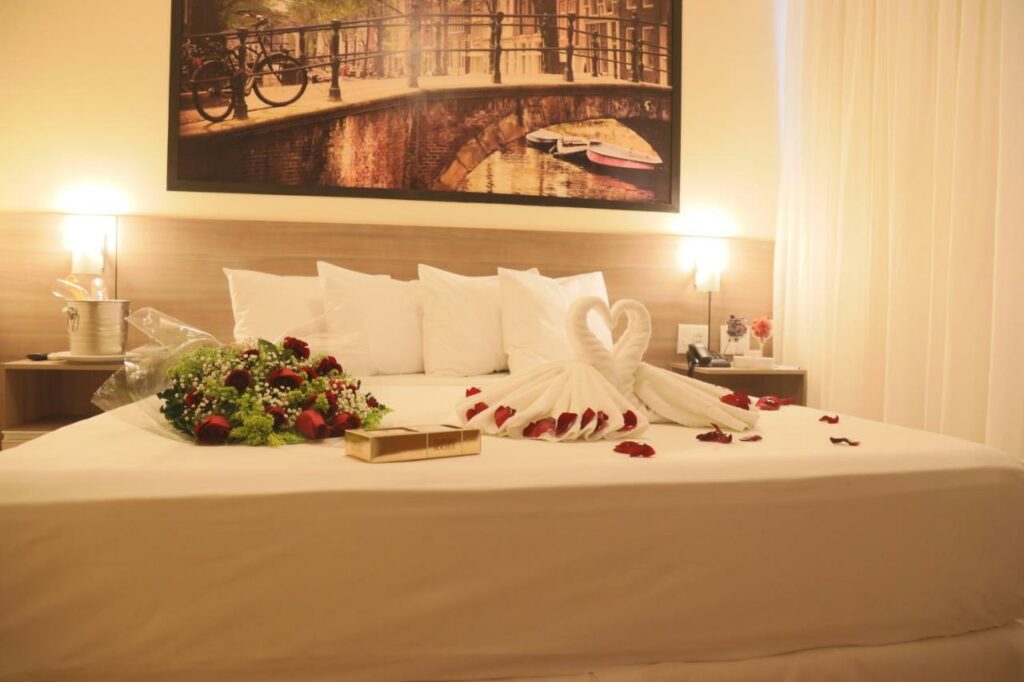 Quarto no Hotel Amsterdam Montes Claros com uma cama de casal decorado com flores e bombons, várias almofadas, dois abajures e um quadro, tudo em tons de branco