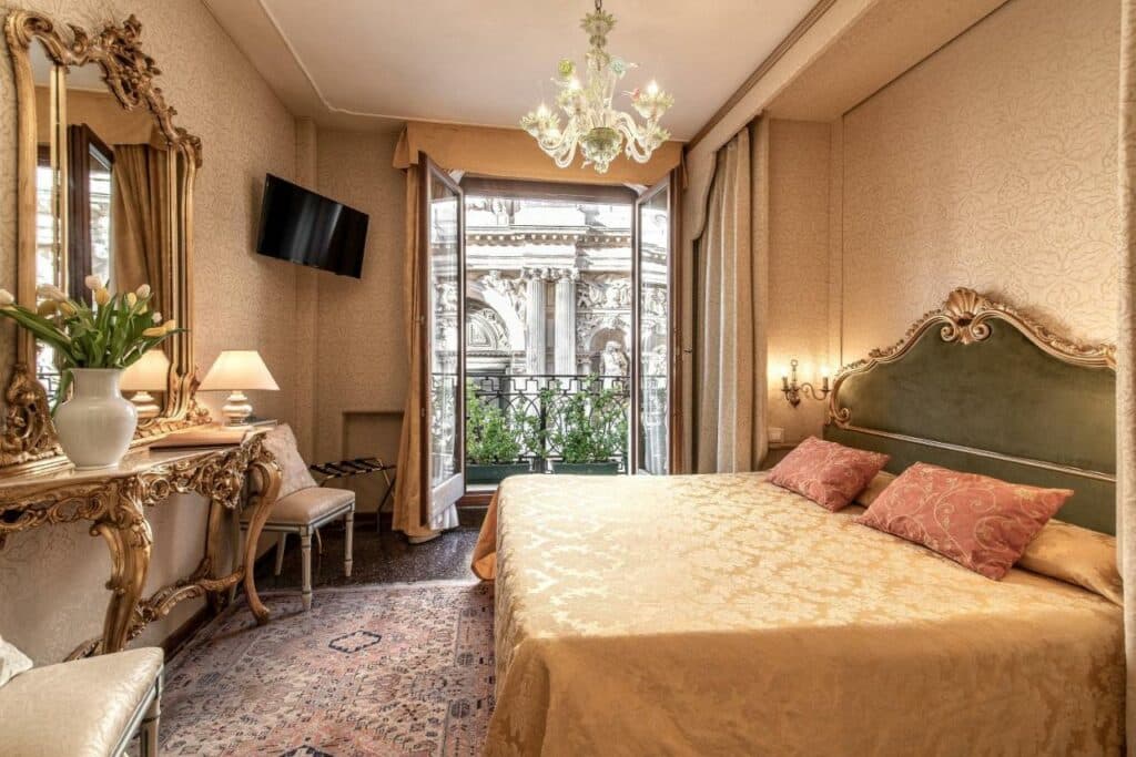 Quarto do Hotel Bel Sito e Berlino com uma sacada com vista para a cidade, uma cama de casal, um espelho, duas poltronas, uma televisão, um abajur e um lustre, tudo em tons de bege e dourado, para representar hotéis em Veneza