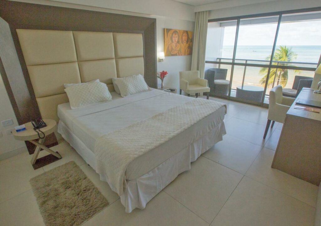 Quarto no Hotel Cabo Branco Atlântico com uma cama de casal, uma mesa com uma poltrona, um quadro, uma sacada ampla com duas poltronas e com vista para o mar, tudo em tons de branco creme e bege, para representar resorts em João Pessoa