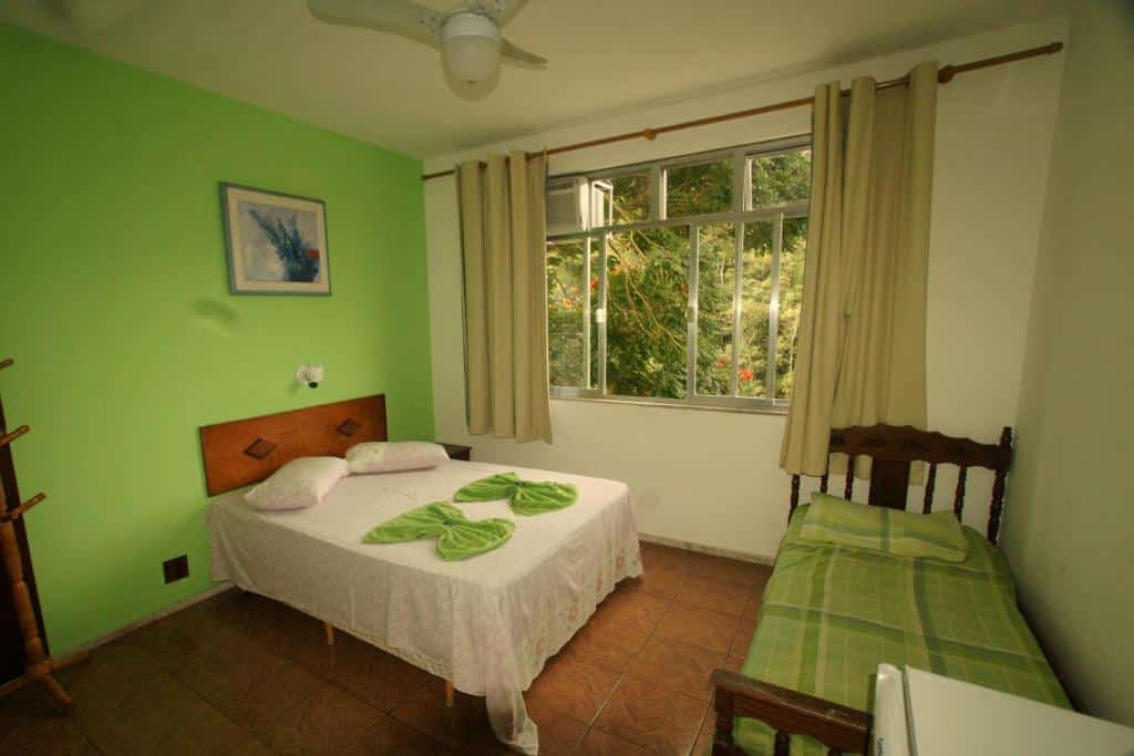 Quarto no Hotel Conora com uma janela dando vista para a natureza, uma cama de casal e uma de solteiro, tudo decorado em tons de verde claro e branco