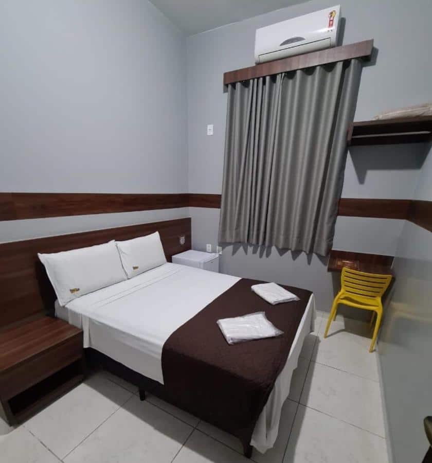 Quarto no Hotel Cerrado com uma cama de casal, uma janela com cortinas, uma pequena mesa com uma cadeira, um frigobar e uma pequena mesinha de cabeceira