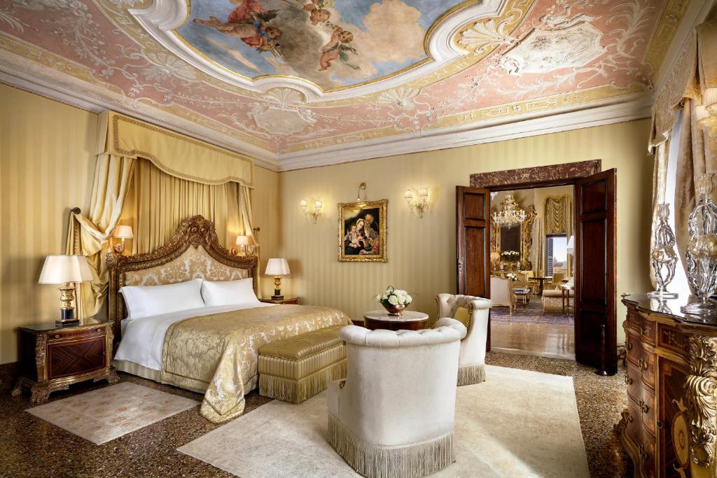 Quarto no Hotel Danieli com tudo decorado em estilo renascentista, em tons de bege e muitos ornamentos na cabeceira da cama de casal, espaço amplo, duas poltronas, uma cômoda, um quadro e dois abajures, para representar hotéis em Veneza
