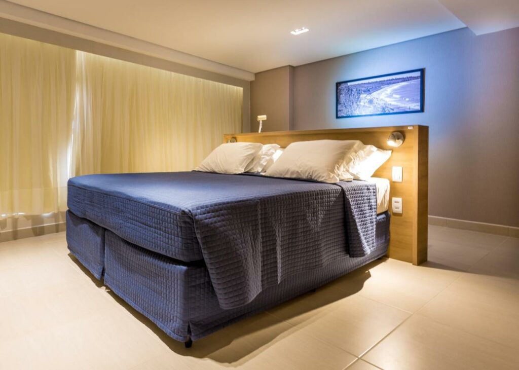 Quarto do Hotel Manaíra com uma cama de casal, um quadro luminoso, uma varanda com cortinas e dois abajures internos na cabeceira, para representar resorts em João Pessoa