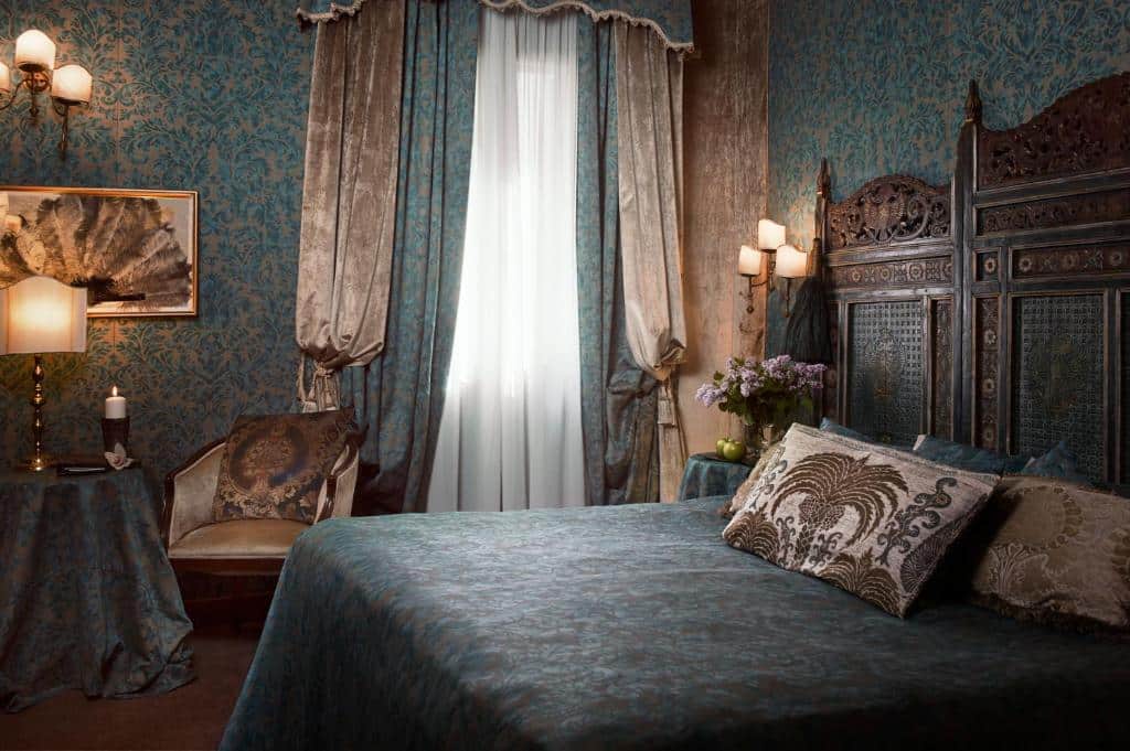 Quarto no Hotel Metropole Venezia em tons de azul e bege, tudo decorado com estilo renascentista e estampas, uma cama de casal com travesseiros, uma mesa com uma abajur e uma poltrona, além da janela com enormes cortinas