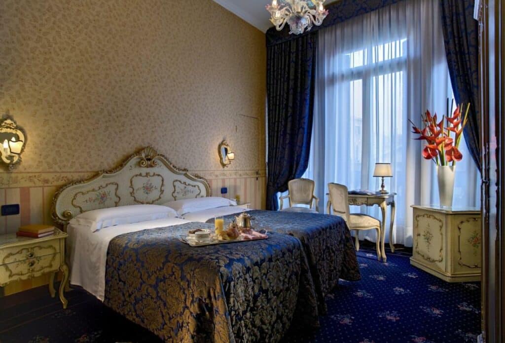 Quarto amplo no Hotel Montecarlo com uma varanda, uma mesa com duas cadeiras, um vaso de flores vermelhas, uma cama de casal e abajures presos na parede, tudo em tom de azul e dourado, também em estilo renascentista, para representar hotéis em Veneza