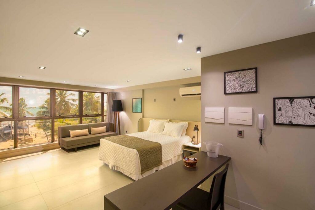 Quarto em Infinity at the Sea by Welkom, um local amplo com janelas dando vista para a praia, uma cama de casal, um ar-condicionado, um pequeno sofá e um abajur